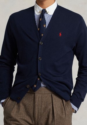 Laivastonsininen neuletakki, jossa punainen kirjailtu logo, laivastonsininen kuvioitu solmio, sinivalkoraidallinen kaulukseen kiinnitettävä paitapusero ja ruskeat ruudulliset housut.