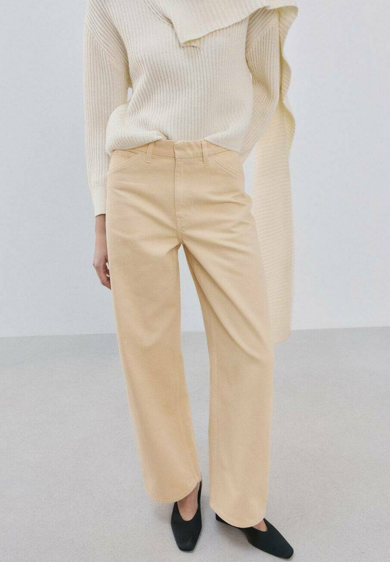 Frau trägt beige weit geschnittene Hosen, cremefarbenen Rippenpullover und schwarze spitze Ballerinas, steht auf einem hellen Boden vor einem schlichten Hintergrund.