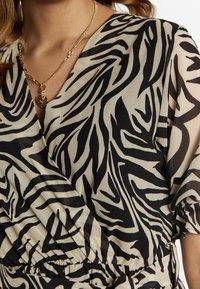 Wickelkleid aus beigem Stoff mit schwarzen Zebrastrifen, tiefem V-Ausschnitt, kurzen Ärmeln und einem gerafften Taillendetail. Akzente durch eine goldene Halskette.