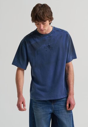 Jeune homme aux cheveux châtain moyen portant un t-shirt ample à manches courtes bleu foncé et un jean bleu, regardant vers le bas avec un tatouage visible sur le bras.