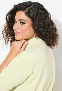 Svetle zelená polo košile s límečkem a krátkými rukávy, měkká textura. Model má kudrnaté vlasy a nosí kulatý zlatý náušník.