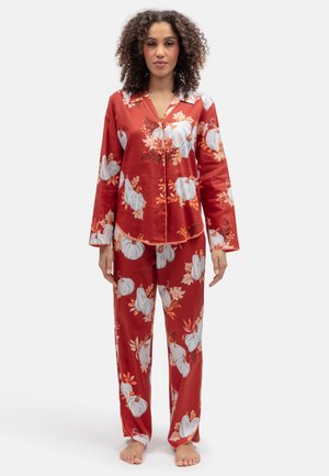 Vrouw draagt rood satijnen pyjamaset met wit pompoen- en oranje bloemmotief, staat blootsvoets op een witte achtergrond.