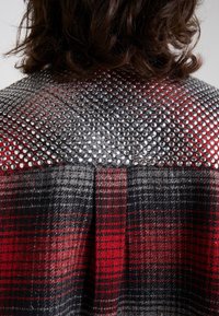 Cheveux noirs jusqu'aux épaules, reposant sur un manteau à carreaux rouge et noir orné de clous argentés couvrant densément la partie supérieure du dos.