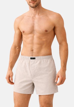 Oberkörperfreier Mann, der hellbeige gestreifte Boxershorts mit elastischem Bund vor einfarbigem Hintergrund trägt.