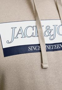 Beige hoodie med et stort hvidt patch med blå tekst "JACK & JONES" og "SINCE NINETEEN." Inkluderer en beige snor. Blød stoftekstur.