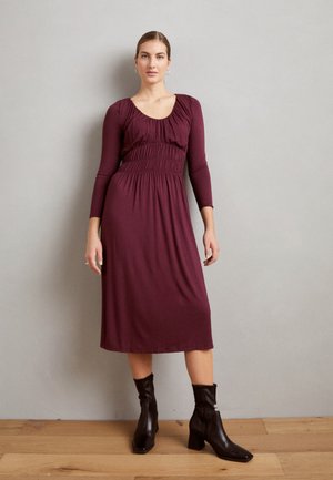 Μπορντό φόρεμα με μακριά μανίκια και φούσκωμα στη μέση, με μήκος μέχρι το γόνατο και λείο ύφασμα; συνδυασμένο με μαύρες μπότες μέχρι τον αστράγαλο.