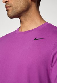 Lilla crew neck t-skjorte laget av mykt stoff, med en svart Nike swoosh-logo på venstre brystområde, jevn tekstur.