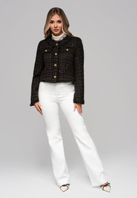 Blonde Frau, die eine schwarz-gold karierte Jacke mit goldenen Knöpfen, einen weißen Rollkragenpullover, weiße Hosen und cremefarbene, spitze Schuhe trägt.