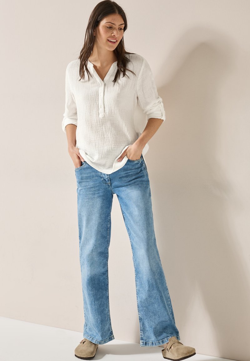 Hvid struktureret bluse med tre knapper ved halsudskæringen, parret med lyseblå flagrende jeans og beige træskoder. En enkel og afslappet påklædning.