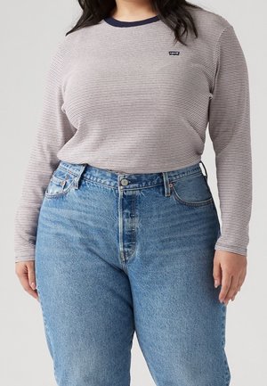 Osoba ubrana w jasnoniebieskie jeansy z wysokim stanem oraz koszulę z długim rękawem w cienkie paski, z ciemnym kołnierzem i małym logo Levi's.