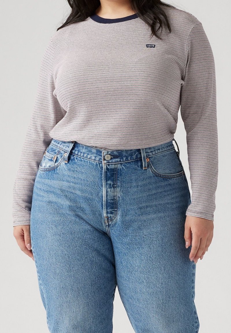 Osoba ubrana w jasnoniebieskie jeansy z wysokim stanem oraz koszulę z długim rękawem w cienkie paski, z ciemnym kołnierzem i małym logo Levi's.