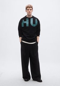 Μαύρο φούτερ με μεγάλο γραφικό "HU" σε τιρκουάζ στο μπροστινό μέρος, συνδυασμένο με φαρδιά μαύρη παντελόνια. Διαθέτει στρογγυλή λαιμόκοψη και μακριά μανίκια.
