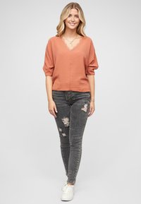 Roodbruin top met een V-hals en gepofte mouwen, gecombineerd met grijze versleten skinny jeans en witte sneakers. Gladde stofstructuur.