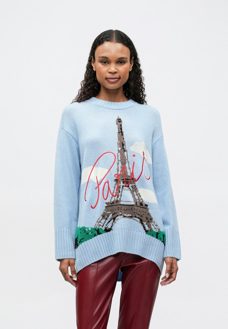 Frau mit lockigem Haar, die einen hellblauen Pullover mit Eiffelturm und dem Text "Paris" trägt, kombiniert mit roten Lederhosen, steht vor einem weißen Hintergrund.