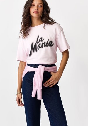 Kobieta z falującymi włosami, ubrana w jasnoróżowy t-shirt "La Mania", ciemnoniebieskie dżinsy i różowy sweter związany wokół talii.