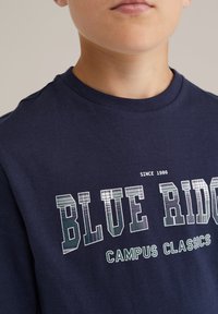 Jūras zila kokvilnas T-krekls ar apaļu kakla izgriezumu, ar baltu un zaļu "BLUE RIDGE CAMPUS CLASSICS" uzrakstu, ar teksturētu grafikas detaļu, vienkāršs dizains.