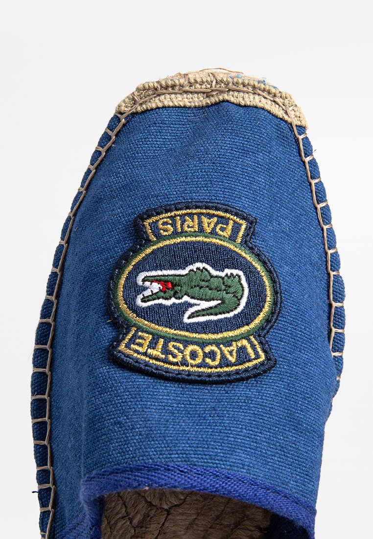 Espadrille bleue avec une tige en toile texturée, présentant un logo crocodile vert brodé et "LACOSTE PARIS" en jaune à l'avant.
