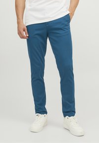 Blå bomullschinos med smal passform, rak fram, och jämn struktur, i kombination med vita sneakers mot en enkel bakgrund.