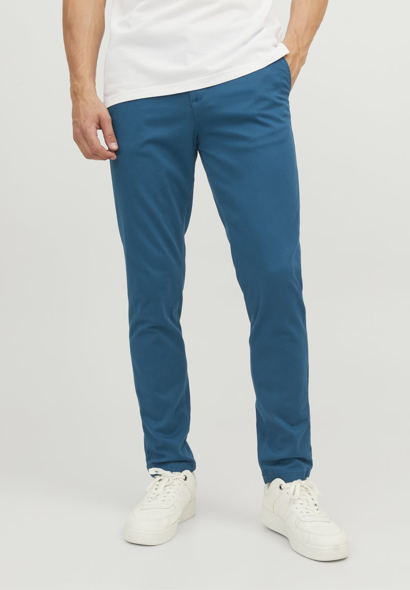 Blå bomullschinos med smal passform, rak fram, och jämn struktur, i kombination med vita sneakers mot en enkel bakgrund.