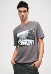 Grå kortærmet t-shirt med et frontalt grafisk print af et sort-hvidt billede, suppleret af en rund hals og blødt stof.