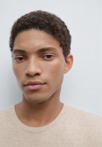 Primo piano di una persona con capelli corti e ricci scuri che indossa una maglietta in maglia beige chiaro, con pelle liscia e sfondo neutro.