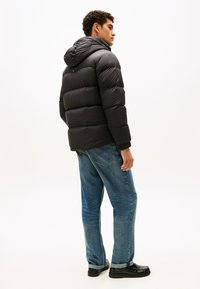 Sort puffjakke med hætte, med quiltede sektioner og elastiske manchetter, båret over blå jeans og sorte sko. Minimalistisk design.
