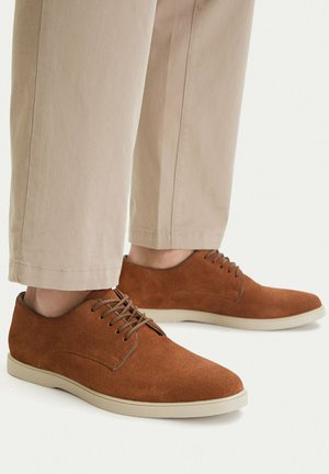 Braune Veloursleder-schnürschuhe getragen mit beigen Hosen, seitliche und vordere Ansichten auf weißem Hintergrund gezeigt.