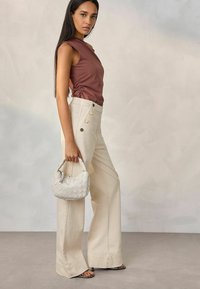 Bruine mouwloze top met rimpelingen, crèmekleurige high-waisted wijde broek met grote knopen, en een kleine witte gevlochten handtas.
