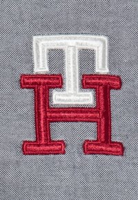 En röd och vit broderad logotyp med ett stiliserat 'H' på en strukturerad grå tygbakgrund. Linjerna är skarpa och väldefinierade.