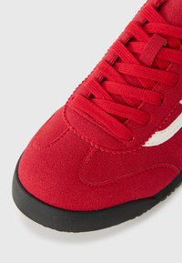 Zbliżenie na czerwony zamszowy przód sneakerów z czerwonymi sznurówkami i czarną gumową podeszwą na białym tle.