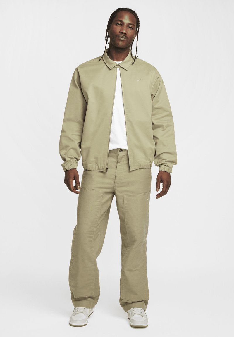 Veste en khaki clair avec col montant et poignets élastiques, associée à un pantalon ample assorti. Portée sur un t-shirt blanc et des baskets blanches.