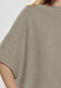 Femme portant un haut en maille côtelée beige avec des manches larges et un col bateau, visible des épaules jusqu'au milieu du bras.