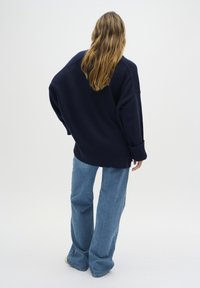 Marineblauwe oversized trui met geribde manchetten en onderrand, gecombineerd met lichtblauwe flared jeans. Zachte gebreide textuur, casual pasvorm en ontspannen silhouet.