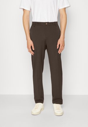 Person trägt dunkelbraune, gerade geschnittene Hosen, weißes T-Shirt und weiße Turnschuhe und steht vor einem einfarbigen Hintergrund.