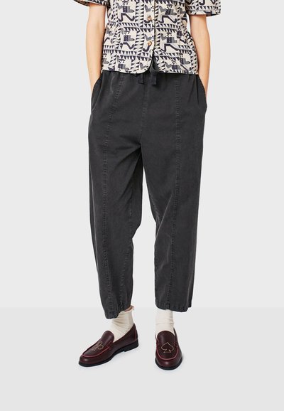 Persoon in losse zwarte broek met elastische enkels, witte sokken en donkergroene instapschoenen met borduursel van kaartpakken.