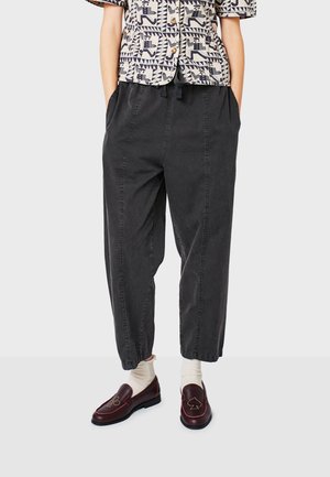 Baggy black cotton trousers with a smooth texture, elasticated cuff, and front pockets; paired with burgundy loafers and cream socks.
