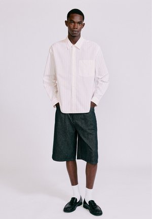 Giovane uomo in piedi che indossa una camicia leggera a righe con maniche lunghe, pantaloncini scuri a righe che arrivano al ginocchio, calzini bianchi e mocassini neri su uno sfondo semplice.
