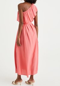 Žena v korálových midi šatech na jedno rameno s volánkovým detailem, stojící, vbeřezovými otevřenými kitten heels na bílém pozadí.
