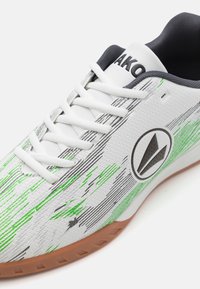 Zapato deportivo blanco con rayas verdes y negras. Presenta una superficie texturizada, lengüeta acolchada y una suela de goma. Logo en el costado.