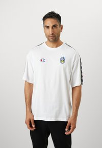 T-shirt en coton blanc avec des manches courtes, arborant un logo de club de football bleu et jaune sur la poitrine et des accents à motifs sur les manches.