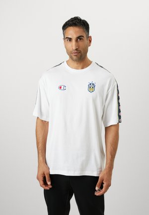 Weißes Baumwoll-T-Shirt mit kurzen Ärmeln, versehen mit einem blauen und gelben Fußballvereinslogo auf der Brust und gemusterten Akzenten an den Ärmeln.