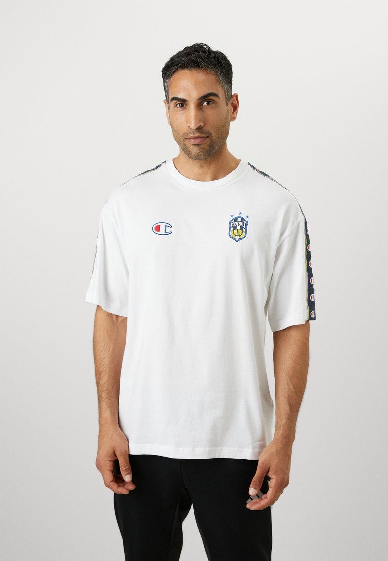T-shirt en coton blanc avec des manches courtes, arborant un logo de club de football bleu et jaune sur la poitrine et des accents à motifs sur les manches.