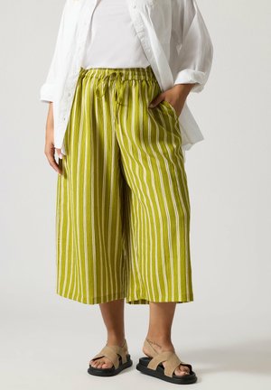 Person trägt weit geschnittene, grün-weiß vertikal gestreifte, gekürzte Hosen mit weitem Bein, ein weißes Hemd und bequeme beigen Sandalen; eine Knöchel-Tätowierung ist sichtbar.