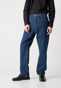 Mørkeblå denimbukser med en afslappet pasform, der har fem lommer og orange syninger, parret med sorte sko og en sort skjorte.