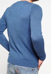 Pull bleu tricoté avec une coupe ajustée, un col rond et des poignets côtelés. La texture est lisse, mettant en valeur un style décontracté.