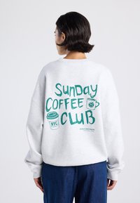 Persona con pelo corto y oscuro que lleva una sudadera blanca con el texto en teal "Club del Café Dominguero" y dibujos de tazas de café en la parte posterior.