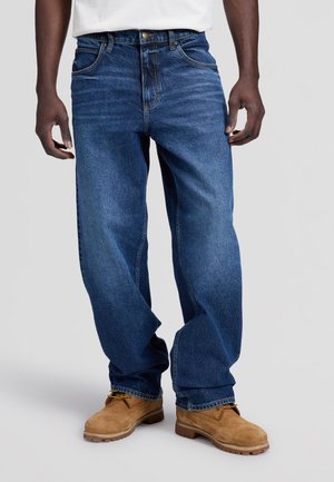 Personne portant un jean bleu ample et des bottes de travail beige, debout devant un fond clair uni.