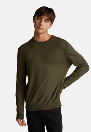 Maglione a maniche lunghe verde oliva in un fine tessuto a coste, con colletto a girocollo e polsini aderenti, indossato con jeans scuri.