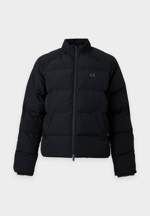 Hombre con gorra negra, chaqueta acolchada negra, suéter beige, pantalones negros y mocasines marrón oscuro con borlas, de pie frente a fondo blanco.