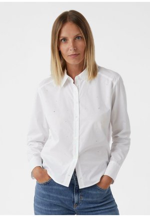 White button-up shirt with subtle embellishments, featuring long sleeves, a collar, and a classic fit. Worn with blue jeans.
